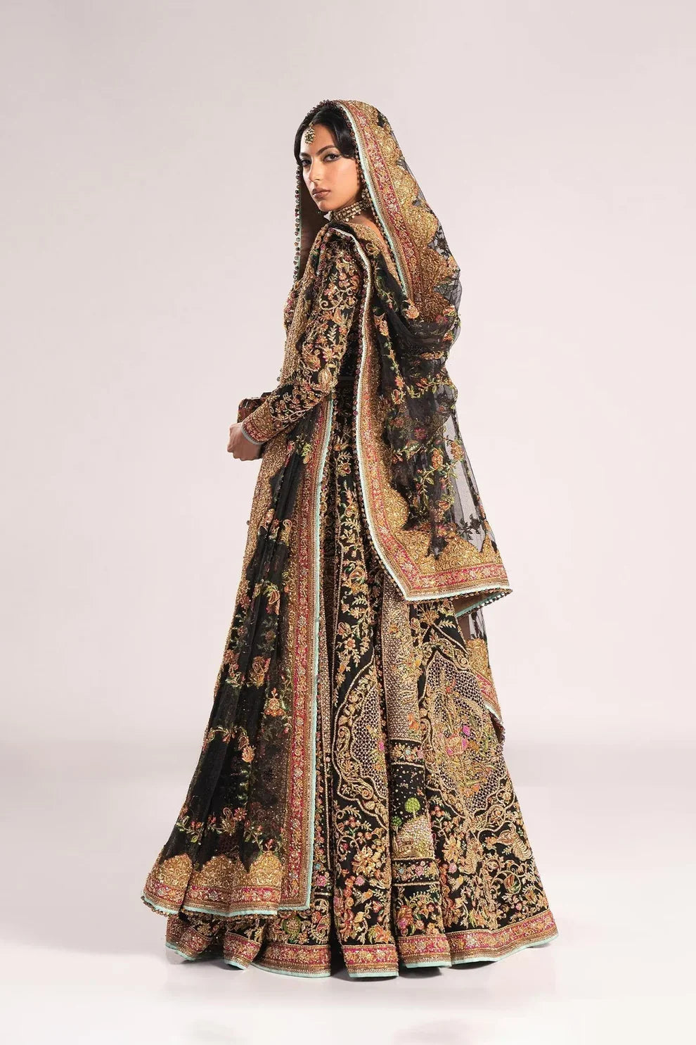 Ajooba by Fahad Hussayn luxury black bridal couture outfit front view featuring heavily hand-embellished net lehenga and blouse with intricate resham, zardozi, naqshi, dabka and crystal embroidery forming rich floral Mughal inspired patterns, dramatic structured silhouette with full flare lehenga and detailed choli, paired with sheer organza dupatta scattered with delicate floral motifs and ornate embroidered border finishing, premium high-end bridal ensemble perfect for weddings, receptions etc