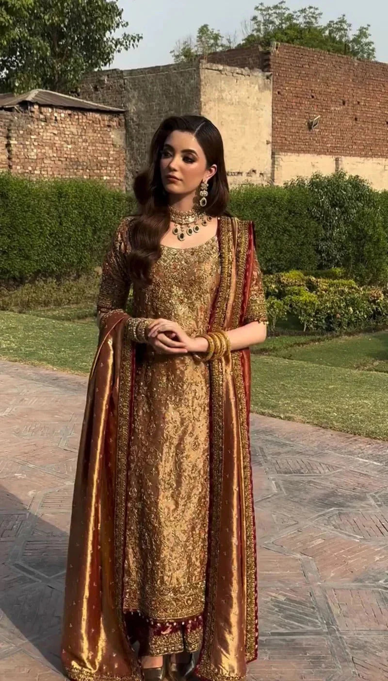 Meerub by Haris Shakeel luxury formal outfit front view featuring deep bronze raw silk shirt with intricate all-over gold hand embroidery, delicate floral motifs, zari and resham threadwork with refined sequin accents, elegantly structured straight silhouette with richly embellished neckline and sleeve borders, paired with matching straight trousers and beautifully coordinated dupatta with embroidered pallu and subtle maroon piping, premium Pakistani designer outfit perfect for weddings, mehndi, baraat etc